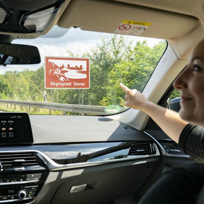 Ein Mann und eine Frau fahren auf der Autobahn. Sie zeigt auf ein braunes Schild mit der Aufschrift "Bergringstadt Teterow"