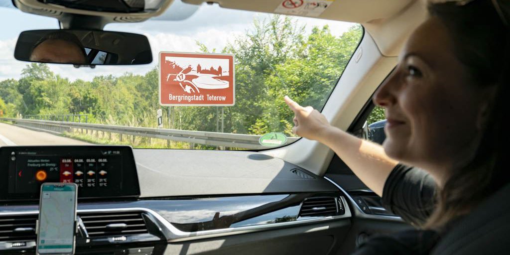 Ein Mann und eine Frau fahren auf der Autobahn. Sie zeigt auf ein braunes Schild mit der Aufschrift "Bergringstadt Teterow"